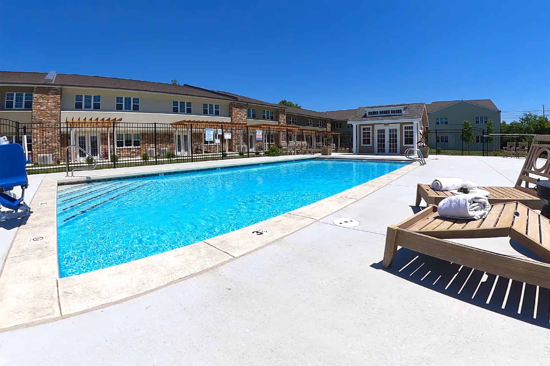 Outdoor swimming pool with lounge chairs and towels in front of a two-story senior living building under a clear blue sky.