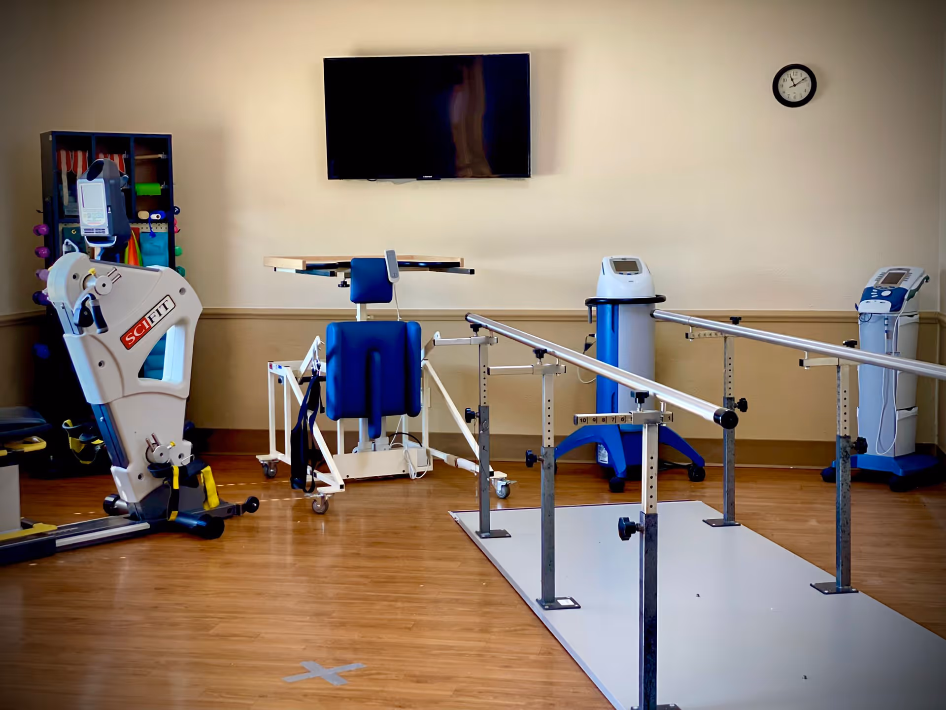 Physical therapy room with parallel bars for walking exercises, exercise equipment including a SciFit machine, a blue padded chair with attached table, and two medical devices on stands. A flat-screen TV is mounted on the beige wall above the equipment, and a clock is visible on the right side of the wall.