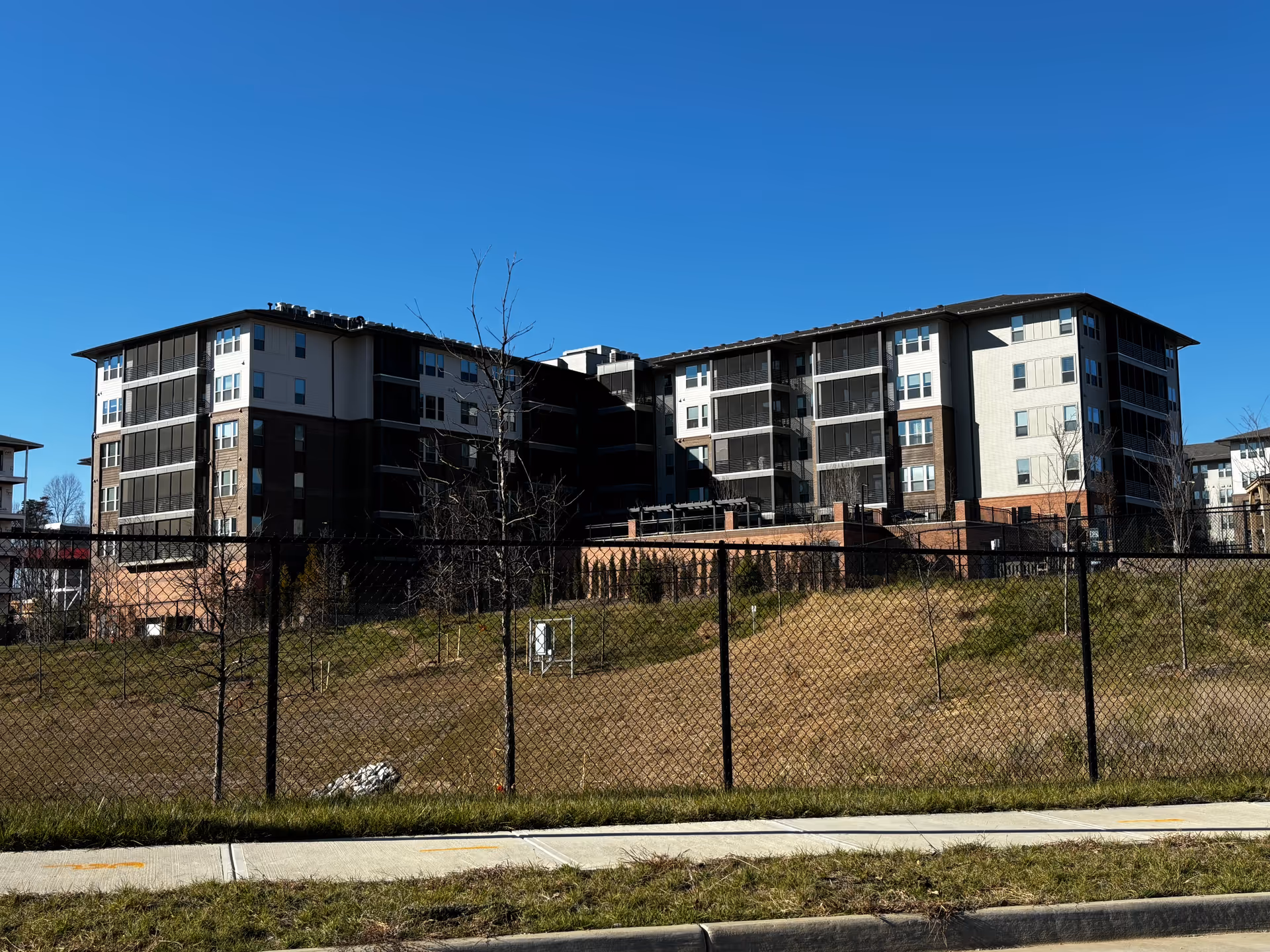 Exterior view of a multi-story senior living facility building under a clear blue sky, with a fenced grassy area in the foreground and a sidewalk along the street.