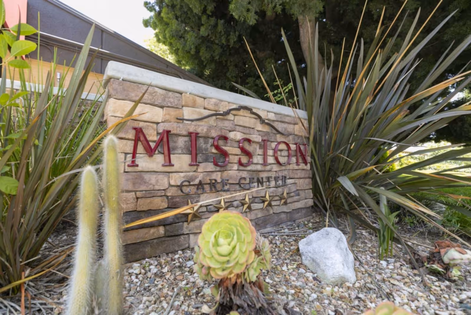 Stone sign for Mission Care Center surrounded by plants and landscaping rocks, with a building and trees in the background.