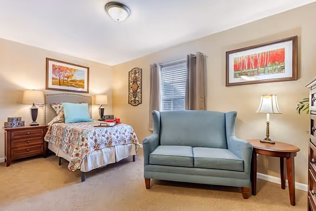A cozy bedroom in a senior living facility featuring a single bed with floral bedding, two bedside tables with lamps, a blue loveseat sofa, a window with gray curtains, and framed artwork on the beige walls.