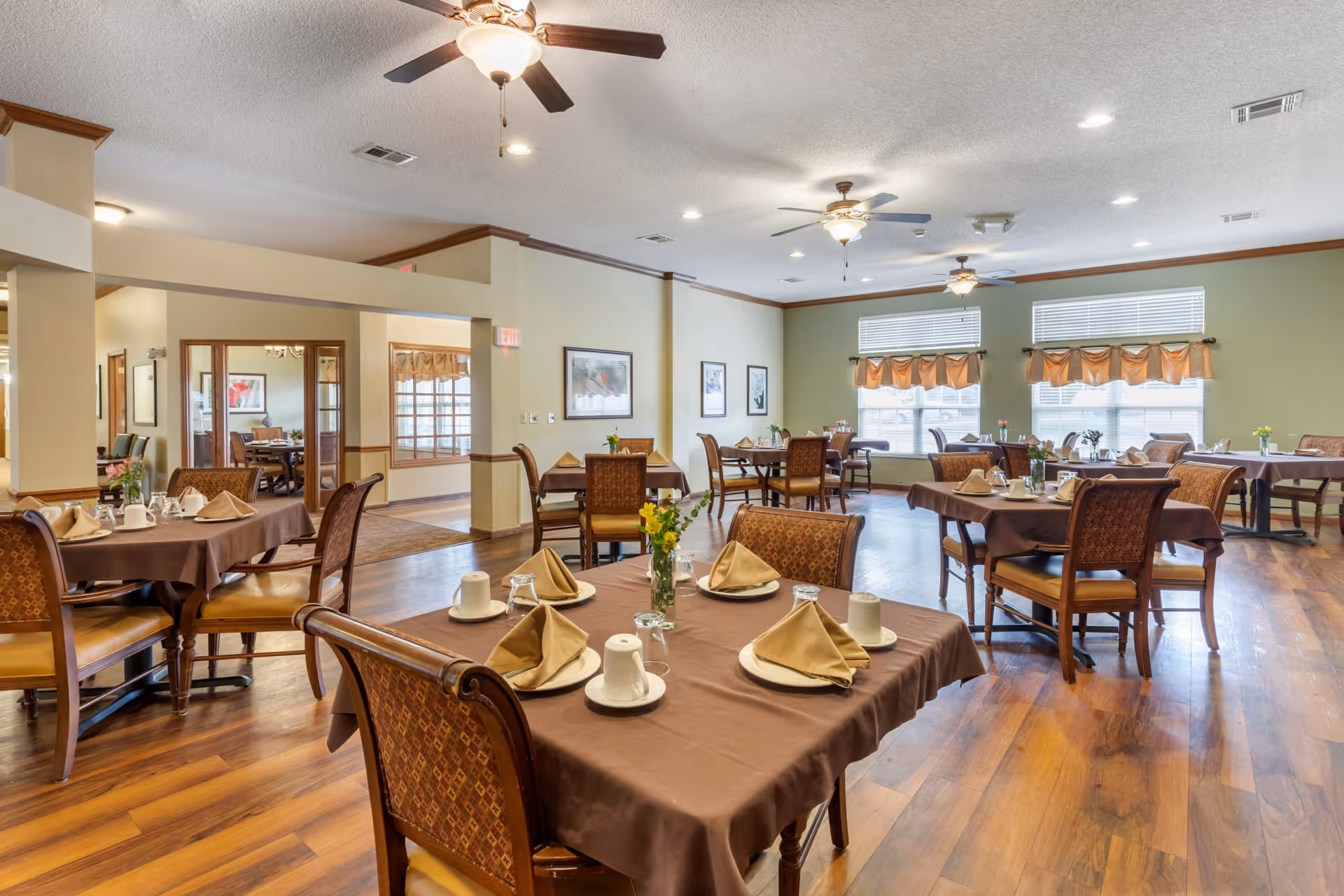 A spacious dining room with multiple tables covered in brown tablecloths, each set with beige napkins, white cups, plates, and glassware. The room has wooden flooring, ceiling fans with lights, and large windows with orange valances allowing natural light to fill the space. There are framed pictures on the walls and a doorway leading to another room.