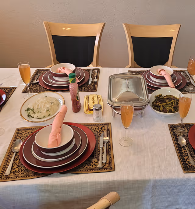A dining table laid out with stacked plates, folded pink napkins, Champagne flutes, side dishes and a covered chafing dish in front of wooden chairs.