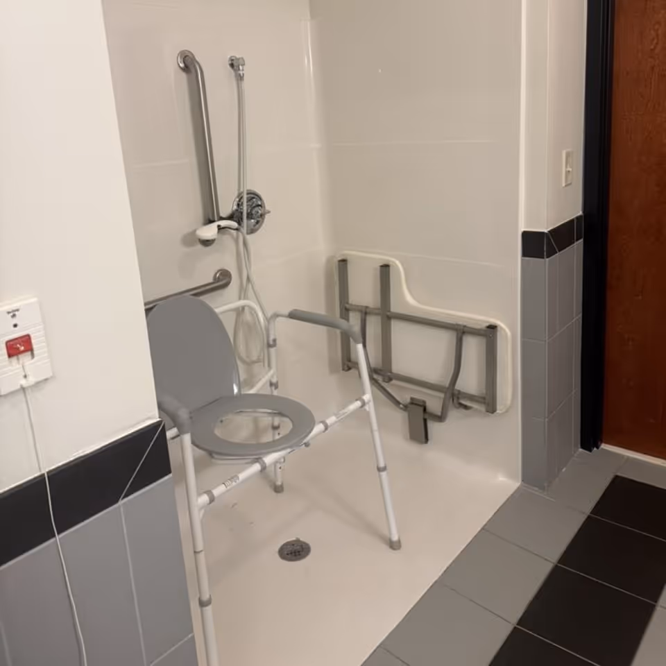 Accessible shower stall with a transfer/commode chair, grab bars, a handheld shower head, and a fold-down bench in a senior facility bathroom.