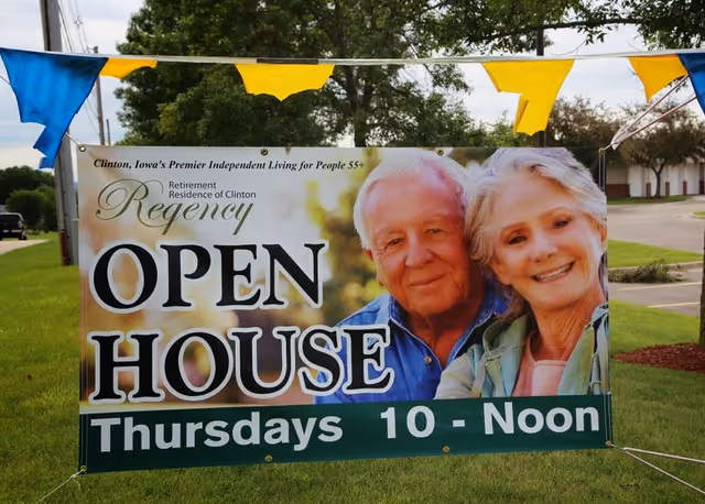 Outdoor banner sign advertising an open house event for Regency Retirement Residence of Clinton, featuring a smiling elderly couple and text indicating the event is on Thursdays from 10 AM to noon. The banner is displayed on a grassy area with trees and a parking lot in the background.