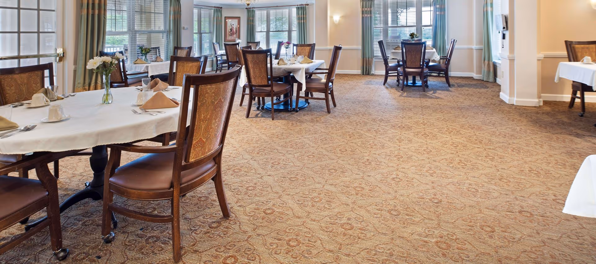 A spacious dining room with multiple tables covered in white tablecloths, each set with cups, glasses, napkins, and silverware. The room has large windows with green curtains allowing natural light to fill the space. The carpet has a patterned design, and the chairs have wooden frames with cushioned seats and backs.
