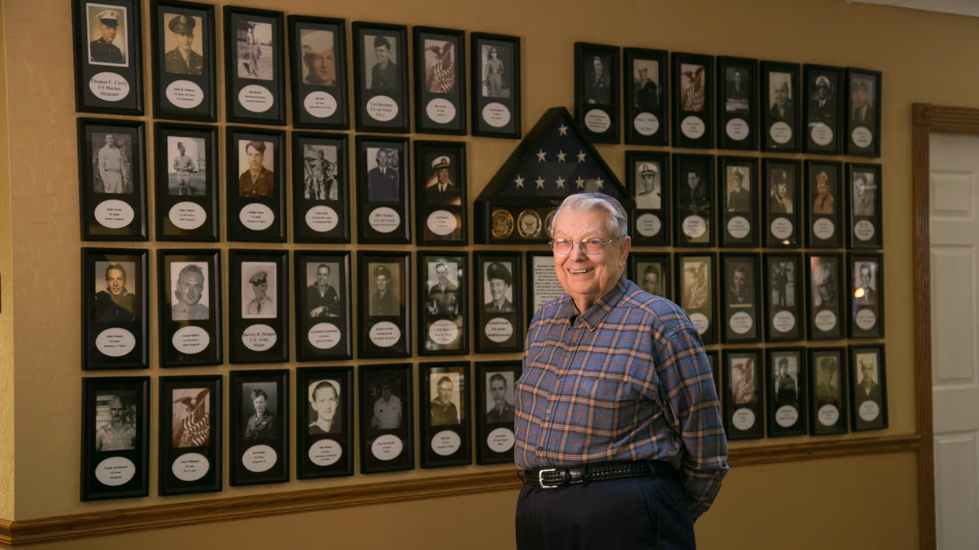 An elderly man wearing glasses and a plaid shirt stands smiling in front of a wall displaying numerous framed black and white photographs of military personnel, along with a folded American flag and military insignia.