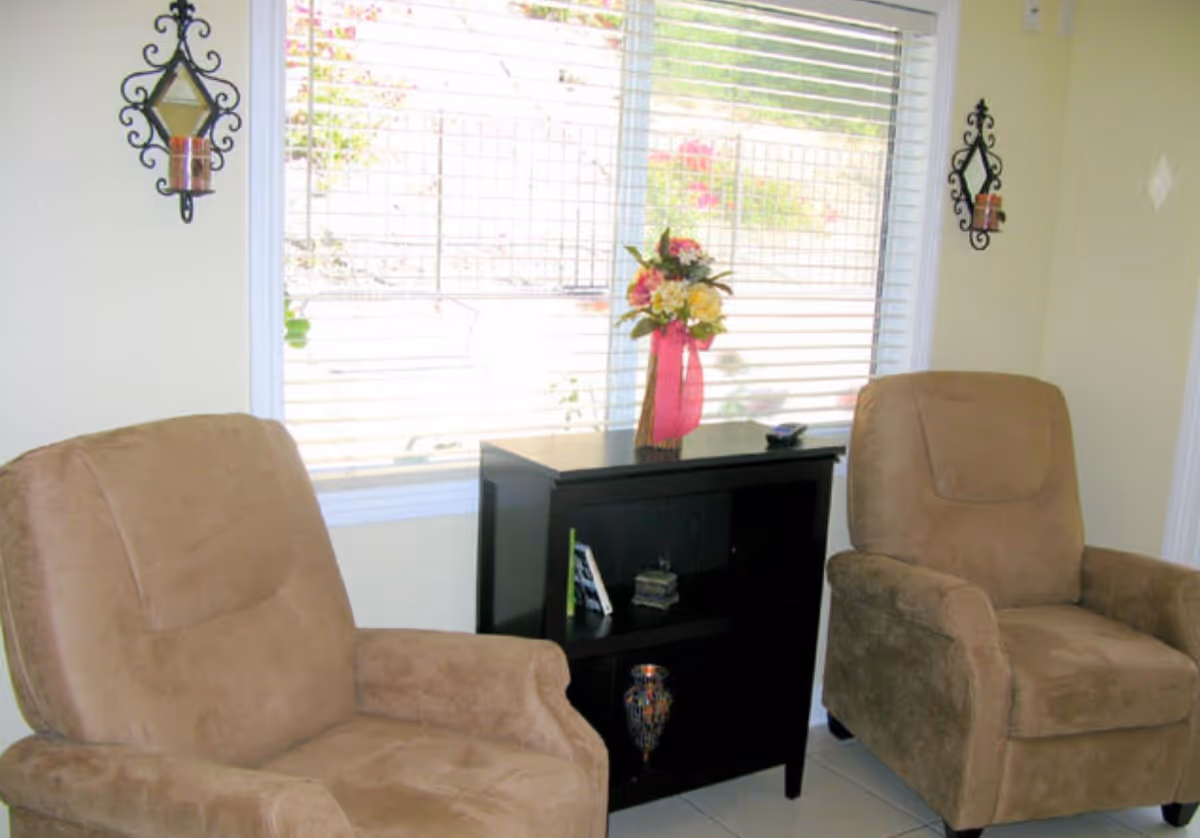 Two beige upholstered armchairs positioned on either side of a black wooden cabinet with shelves. On top of the cabinet is a vase with a bouquet of flowers tied with a pink ribbon. Behind the cabinet is a large window with white blinds partially open, letting in natural light. The walls are light-colored with decorative black metal candle holders mounted on either side of the window.