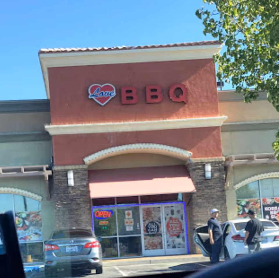 Exterior view of a restaurant named 'Love BBQ' with a red and beige facade, a red awning above the entrance, and two men standing near a white car in the parking lot. The restaurant has an 'OPEN' neon sign and promotional posters on the glass doors.