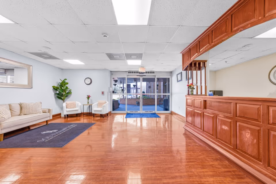 A bright and clean reception area with a wooden front desk on the right, a seating area with a beige couch and two white chairs on the left, a large mirror on the wall, a clock, and a potted plant. The floor is polished wood, and there are glass double doors at the back leading outside.