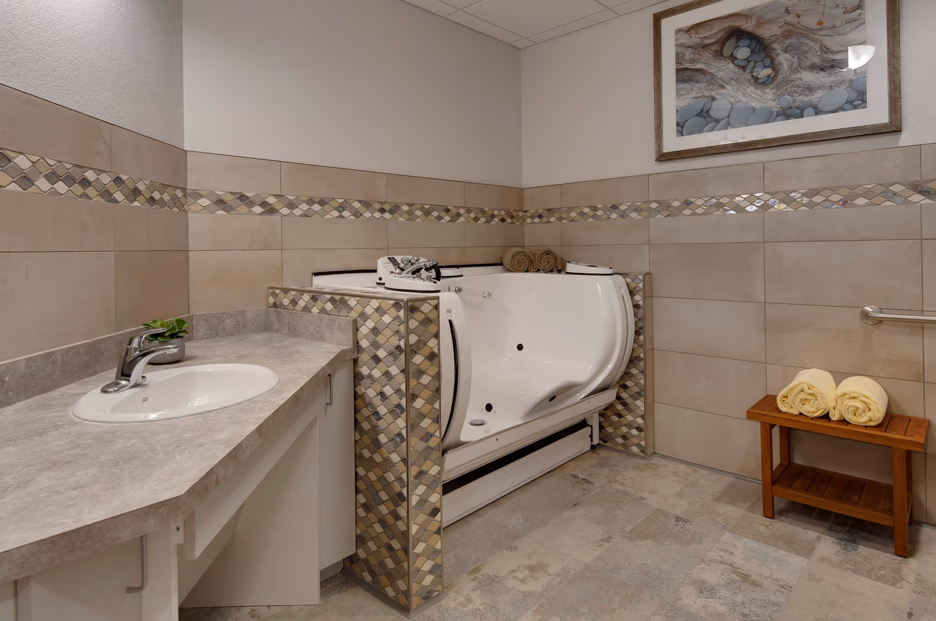 A bathroom with beige tiled walls featuring a decorative mosaic border. There is a white walk-in bathtub with control knobs and two rolled towels on its edge. To the left, a countertop with a sink and a small green plant is visible. On the right, a wooden bench holds two rolled yellow towels. A framed picture of stones and driftwood hangs on the wall above the bathtub.