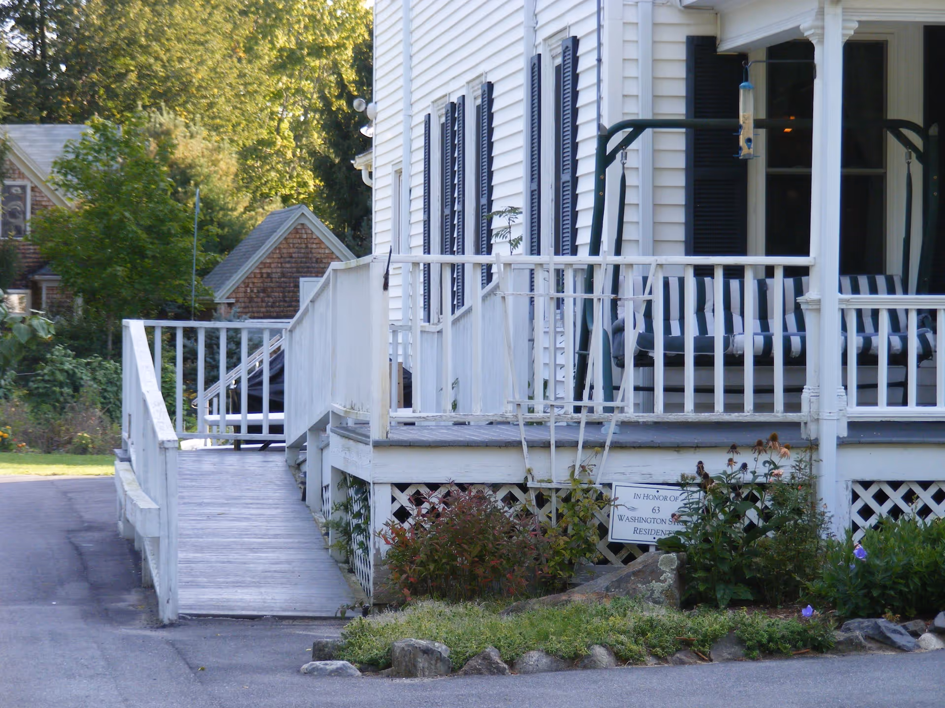 White house front porch with a wooden ramp, striped porch swing cushions, and a small garden bed in front.