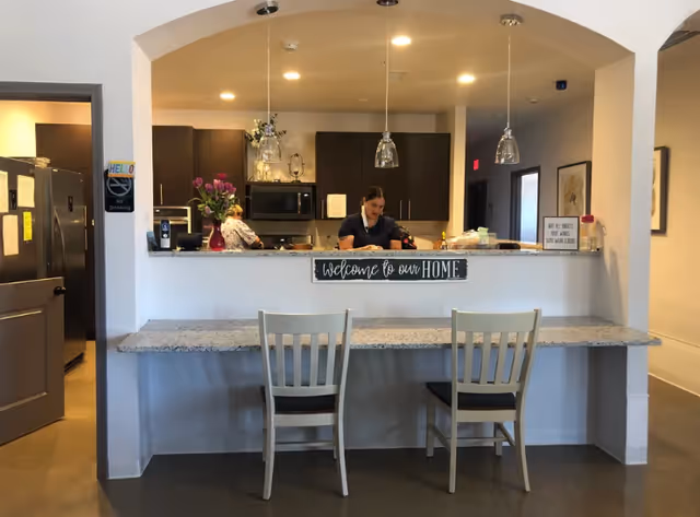 Interior view of a kitchen area in a senior living facility with two women working behind a counter. The counter has a granite surface with two chairs in front. Above the counter, there are three hanging pendant lights and a sign that reads 'Welcome to our HOME'. The kitchen has dark cabinets, a microwave, and various items on the counter. There is a 'No Smoking' sign on the wall near an open door to the left.