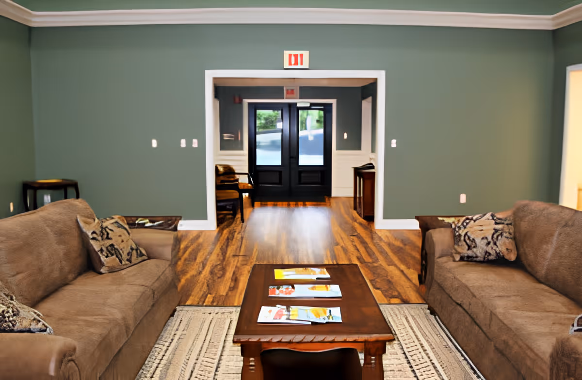 A cozy living room area with two brown sofas facing each other, each with patterned throw pillows. A wooden coffee table with magazines on it sits on a patterned rug between the sofas. The room has green walls, wooden flooring, and a doorway leading to a hallway with double glass doors at the end.