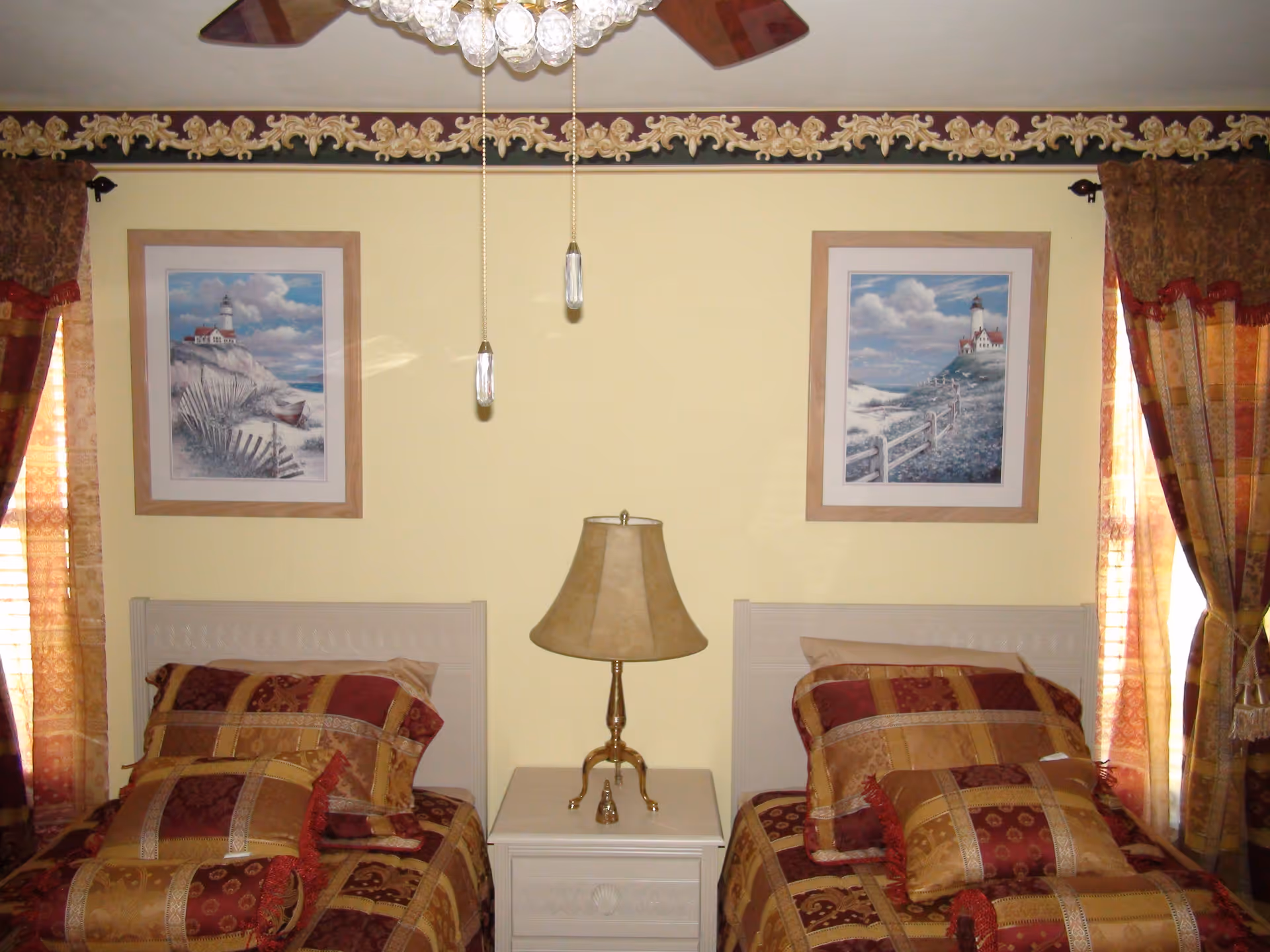 A bedroom with two twin beds separated by a white nightstand with a beige lamp. Each bed has matching patterned bedding in shades of red, gold, and brown. Above each bed hangs a framed picture of a lighthouse scene. The room has yellow walls, decorative border trim near the ceiling, and windows with patterned curtains on either side.