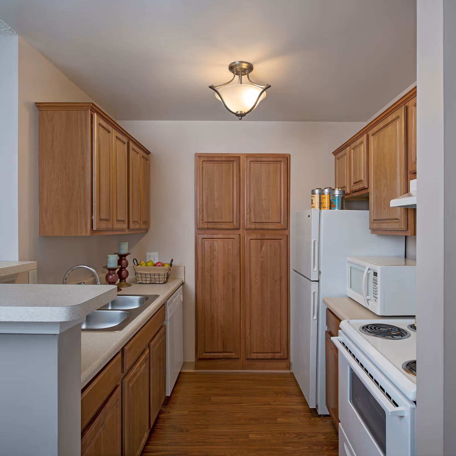 A compact kitchen with wooden cabinets, a white refrigerator, microwave, electric stove, and a double sink. The kitchen has a light fixture on the ceiling and a basket of fruit on the counter.