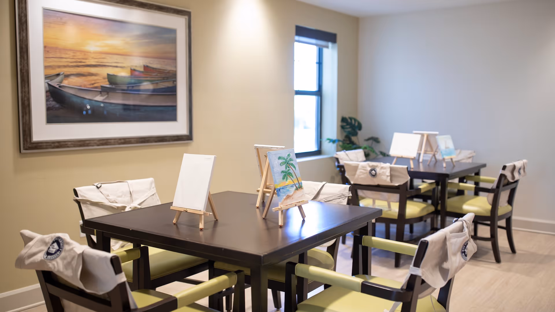 A bright room with two dark wooden tables, each surrounded by four chairs with light green cushions. Small easels with blank and partially painted canvases are placed on the tables. The chairs have white cloth covers draped over their backs. A large framed picture of boats on a beach at sunset hangs on the wall, and a window lets in natural light. There is a potted plant in the corner of the room.