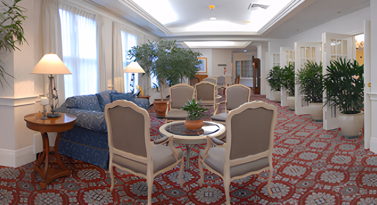 Bright communal lounge with upholstered chairs, round tables, a sofa and many potted plants.