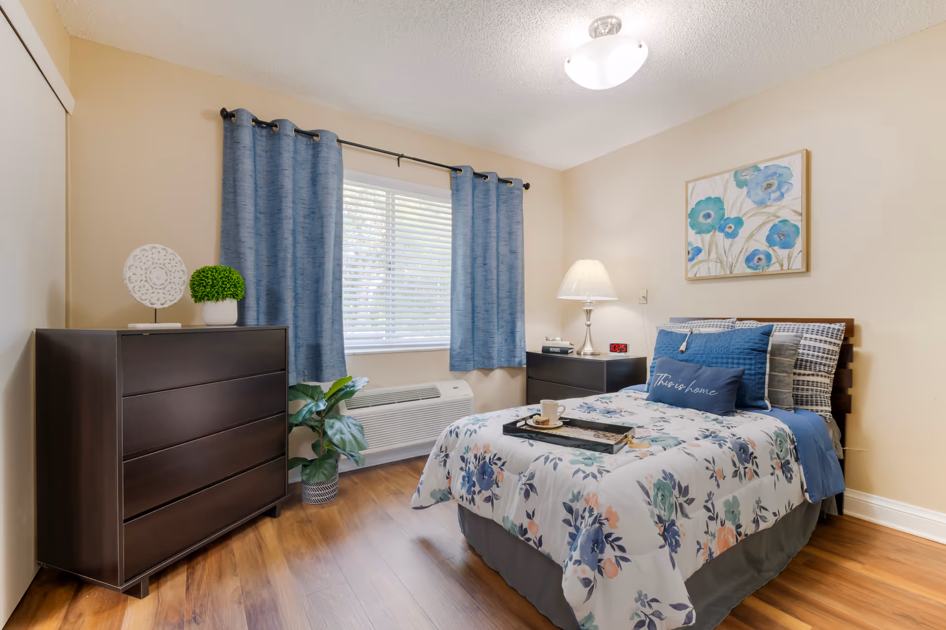 Well-lit furnished bedroom with a floral bed, blue curtains, a dark dresser, bedside table with lamp, and wood flooring.