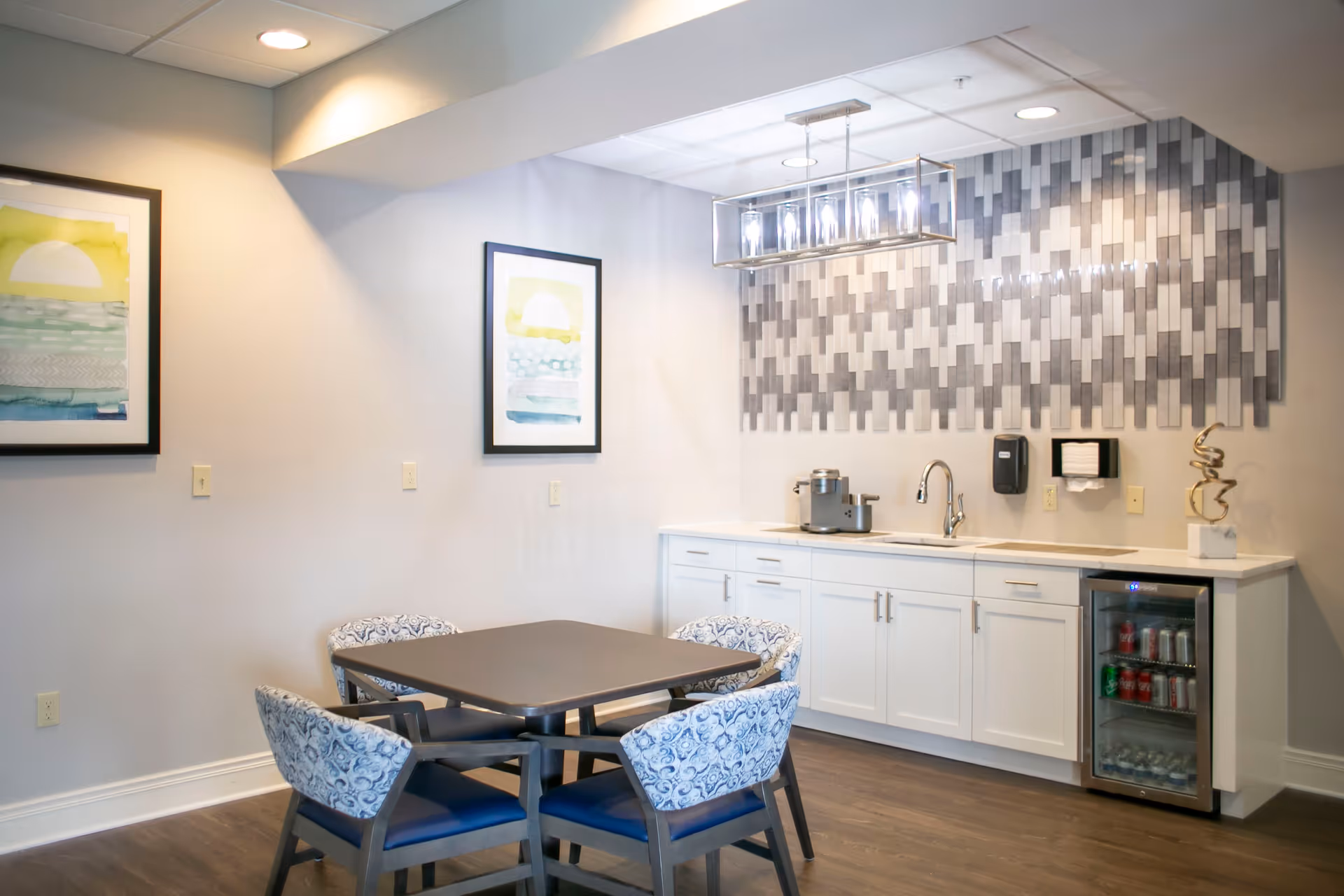A small dining area with a square table surrounded by four patterned chairs. Behind the table is a kitchenette with white cabinets, a sink, a small beverage fridge stocked with drinks, a coffee machine, and a modern light fixture above. The wall behind the kitchenette features a decorative tile backsplash in shades of gray, and two framed abstract paintings hang on the adjacent wall.