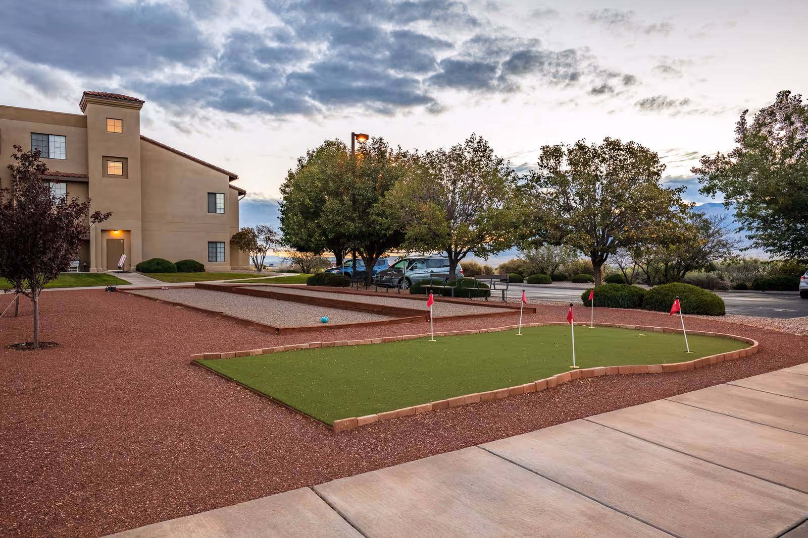 Outdoor area at Fairwinds - Rio Rancho featuring a small putting green with five red flags and a bocce ball court. There are trees, parked cars, and a beige multi-story building in the background under a cloudy sky.