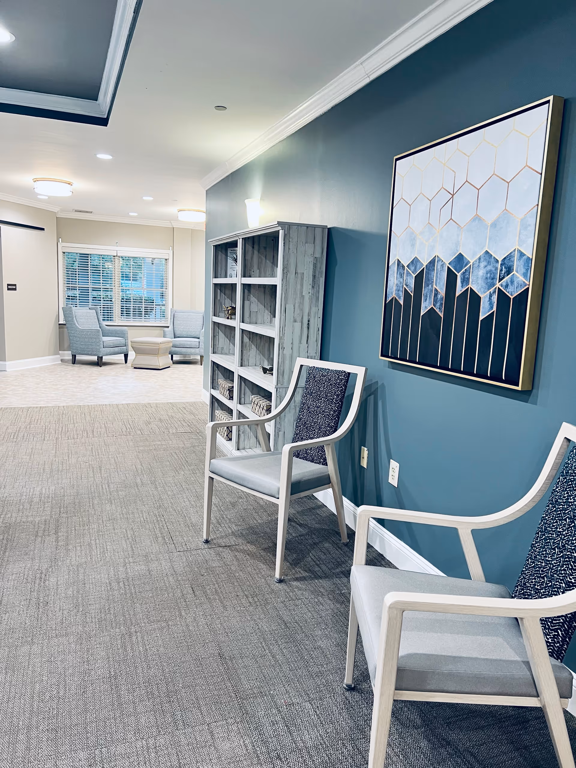 A modern senior living facility interior with two light gray chairs with patterned backs against a teal wall. A large abstract geometric painting in shades of blue and gold hangs above the chairs. Next to the chairs is a tall, open shelving unit with decorative items. In the background, there are two more light gray armchairs and a small ottoman near a window with white blinds. The floor is carpeted in the foreground and tiled in the background, with recessed ceiling lights illuminating the space.