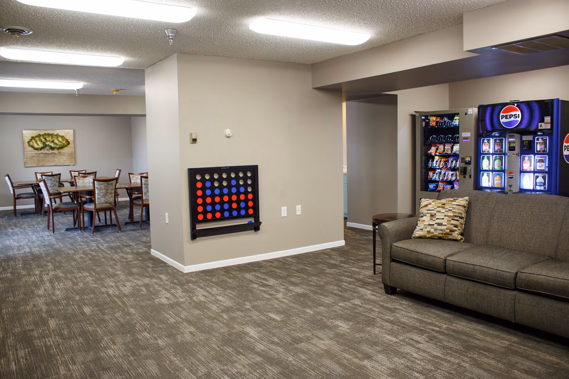 Community lounge with a sofa, vending machines, a wall-mounted Connect Four game and dining tables visible in the background.