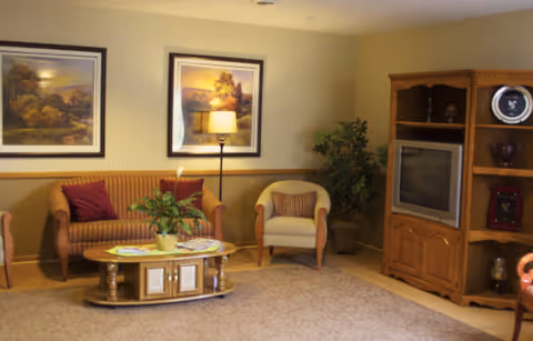 A cozy living room with a striped sofa and two armchairs, a wooden coffee table with a potted plant and magazines, two framed landscape paintings on the wall, a floor lamp, a large wooden entertainment center with a TV and decorative items, and a potted plant in the corner.
