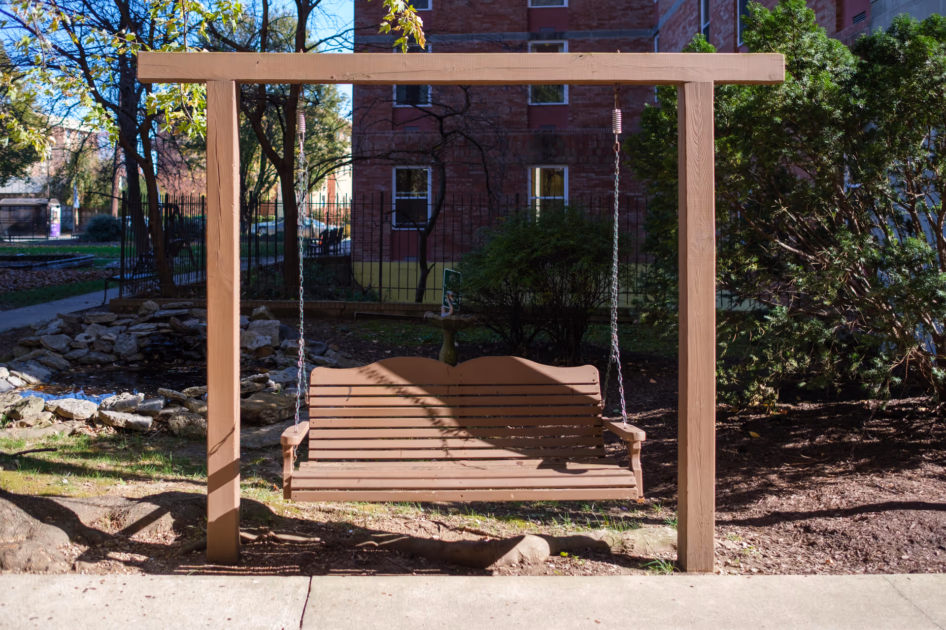 Wooden porch swing hanging from a frame in a landscaped courtyard with a small pond and a brick building in the background.