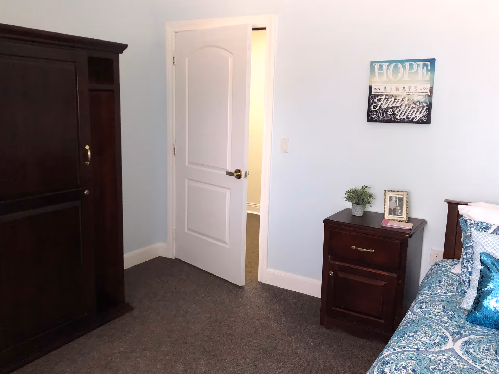 A small bedroom with a partially open white door, dark wood armoire and nightstand beside a bed with blue patterned bedding.