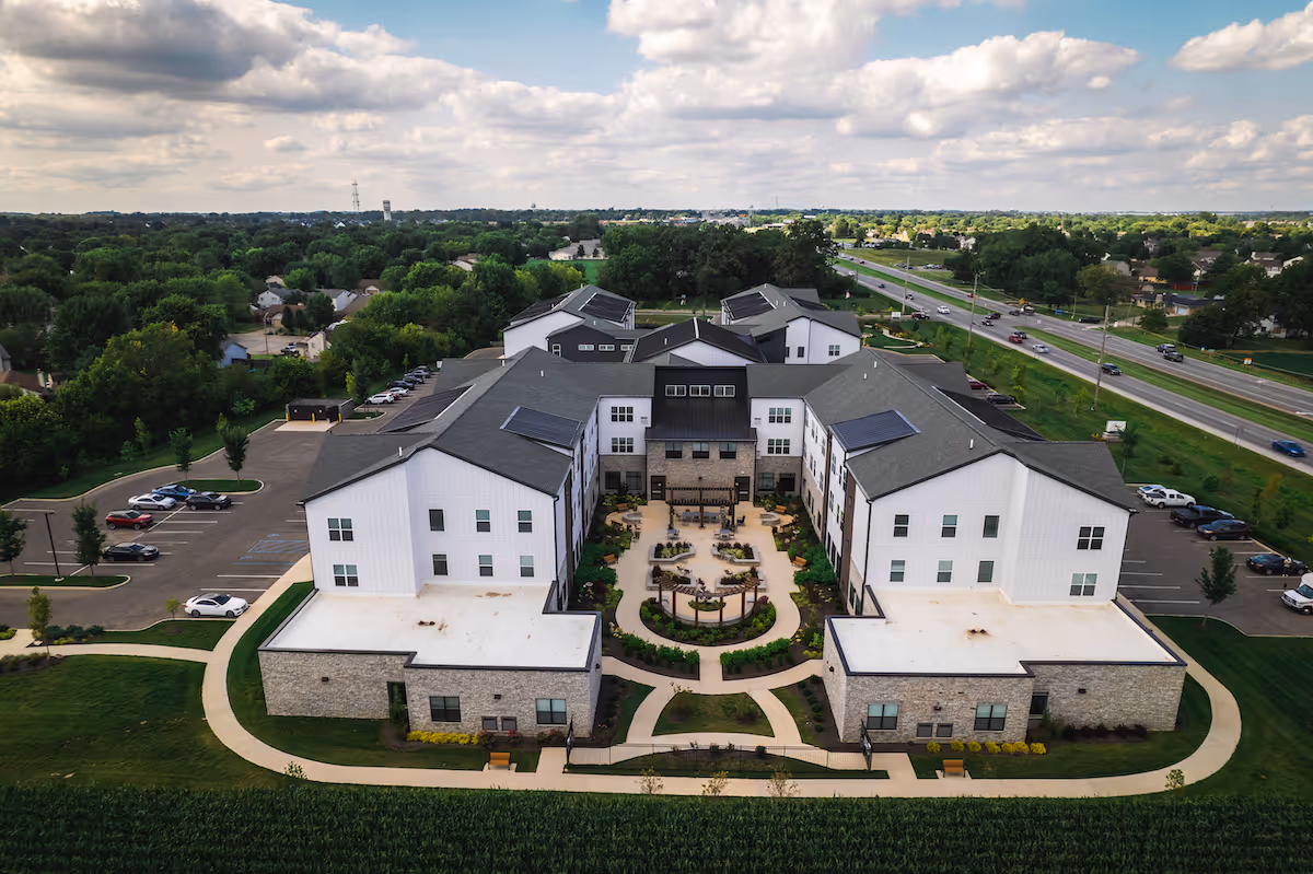 Aerial view of a large senior living facility named Astral at Franklin, featuring a U-shaped building with white and stone exterior walls, surrounded by landscaped gardens, walkways, parking lots, and a nearby road with vehicles. The sky is partly cloudy.