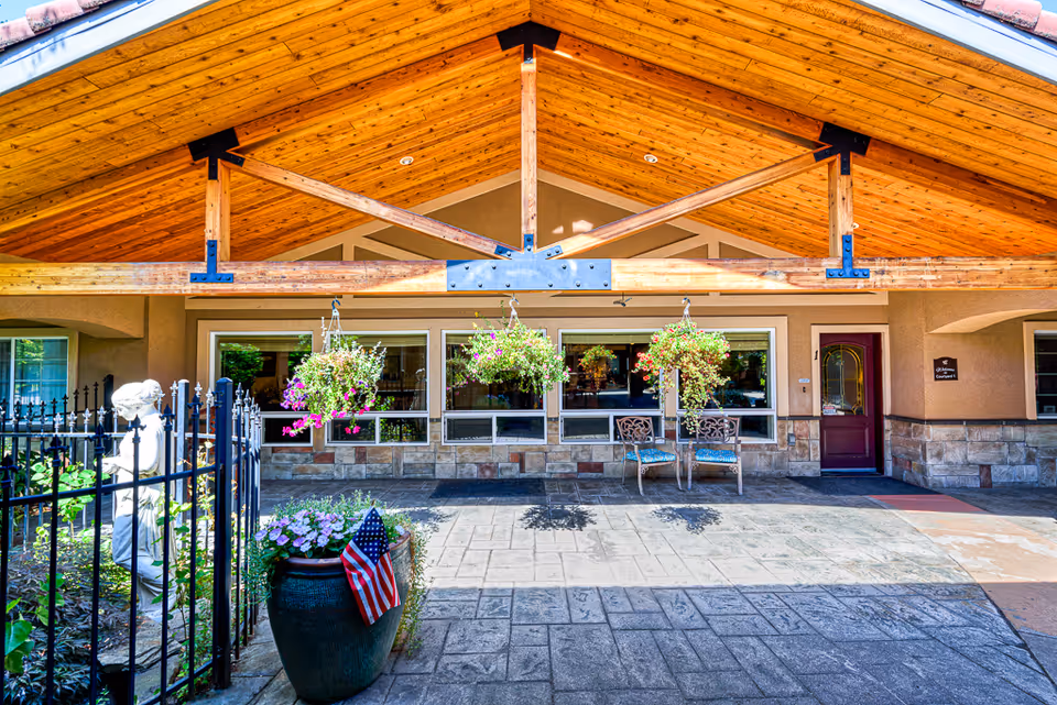 Entrance area of a senior living facility with a wooden peaked roof supported by beams, hanging flower baskets, a large planter with flowers and an American flag, a black metal fence with a white statue behind it, and two chairs near a window next to a red door.