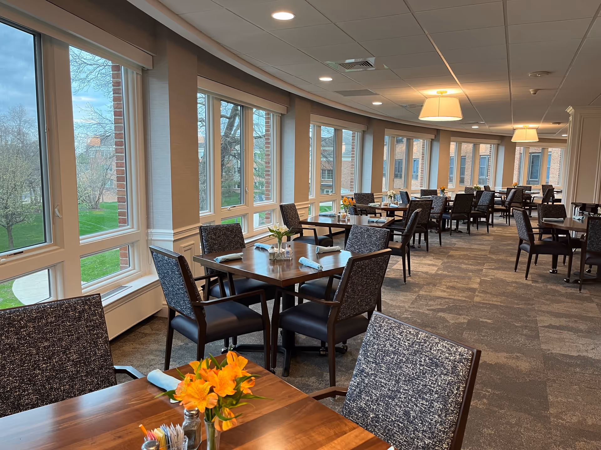 A spacious dining room with multiple wooden tables and cushioned chairs arranged neatly. Each table has a small vase with fresh flowers and rolled napkins. Large windows line one side of the room, allowing natural light to brighten the space and offering a view of green trees and a brick building outside. The ceiling has recessed lighting and hanging lamps.