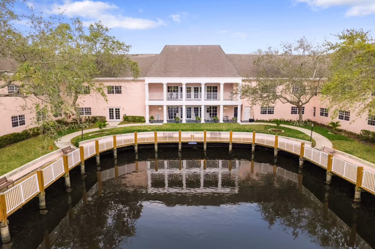 Exterior view of a pink two-story building with a sloped roof, surrounded by trees and a curved wooden railing along a body of water in the foreground. The building has multiple windows and a central balcony area with white columns.