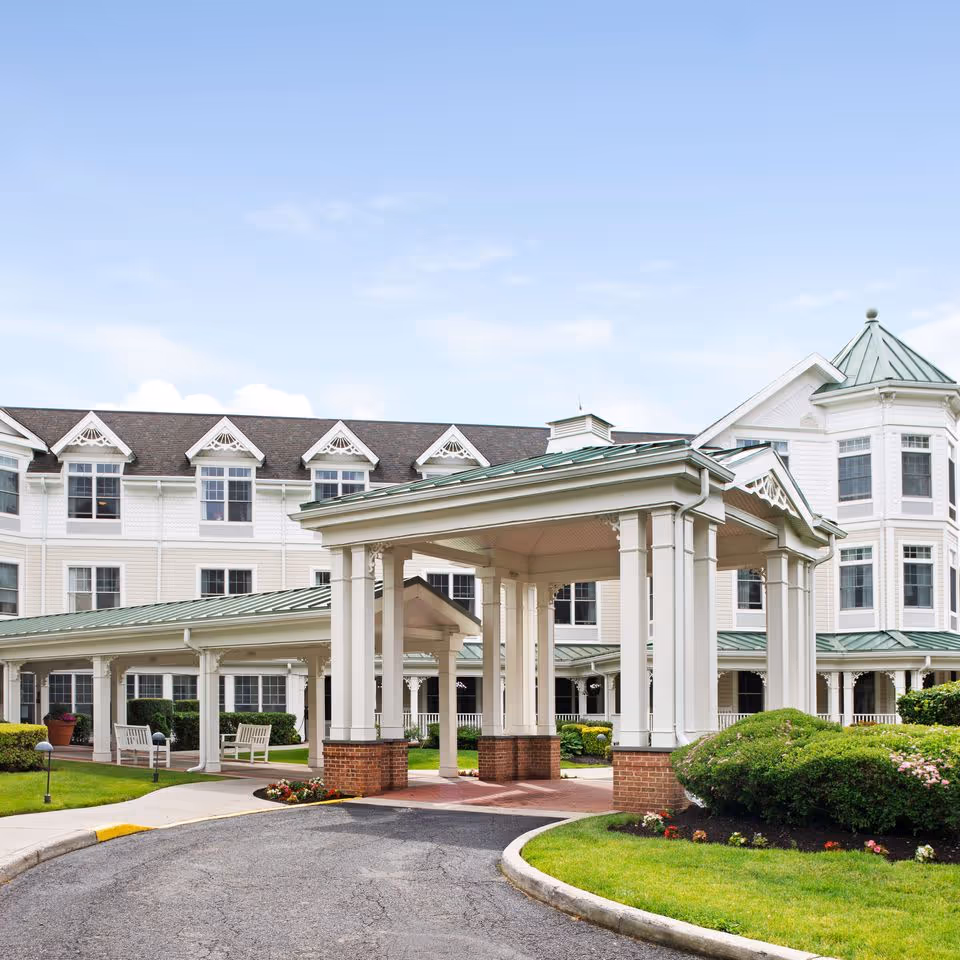 Front entrance and porte-cochère of a white multi-story senior living building with green metal roofs and landscaped grounds.