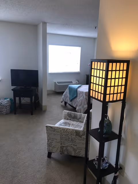 Small studio bedroom with a bed by the window, TV on a stand, patterned armchair, and an illuminated tall floor lamp.