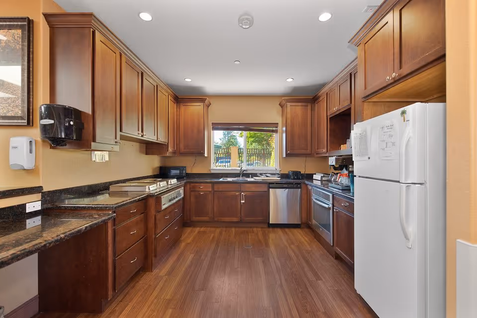 A spacious kitchen with wooden cabinets, granite countertops, and hardwood flooring. The kitchen features a white refrigerator, stainless steel dishwasher, oven, and stove. There is a window above the sink letting in natural light and a paper towel dispenser and soap dispenser mounted on the wall.