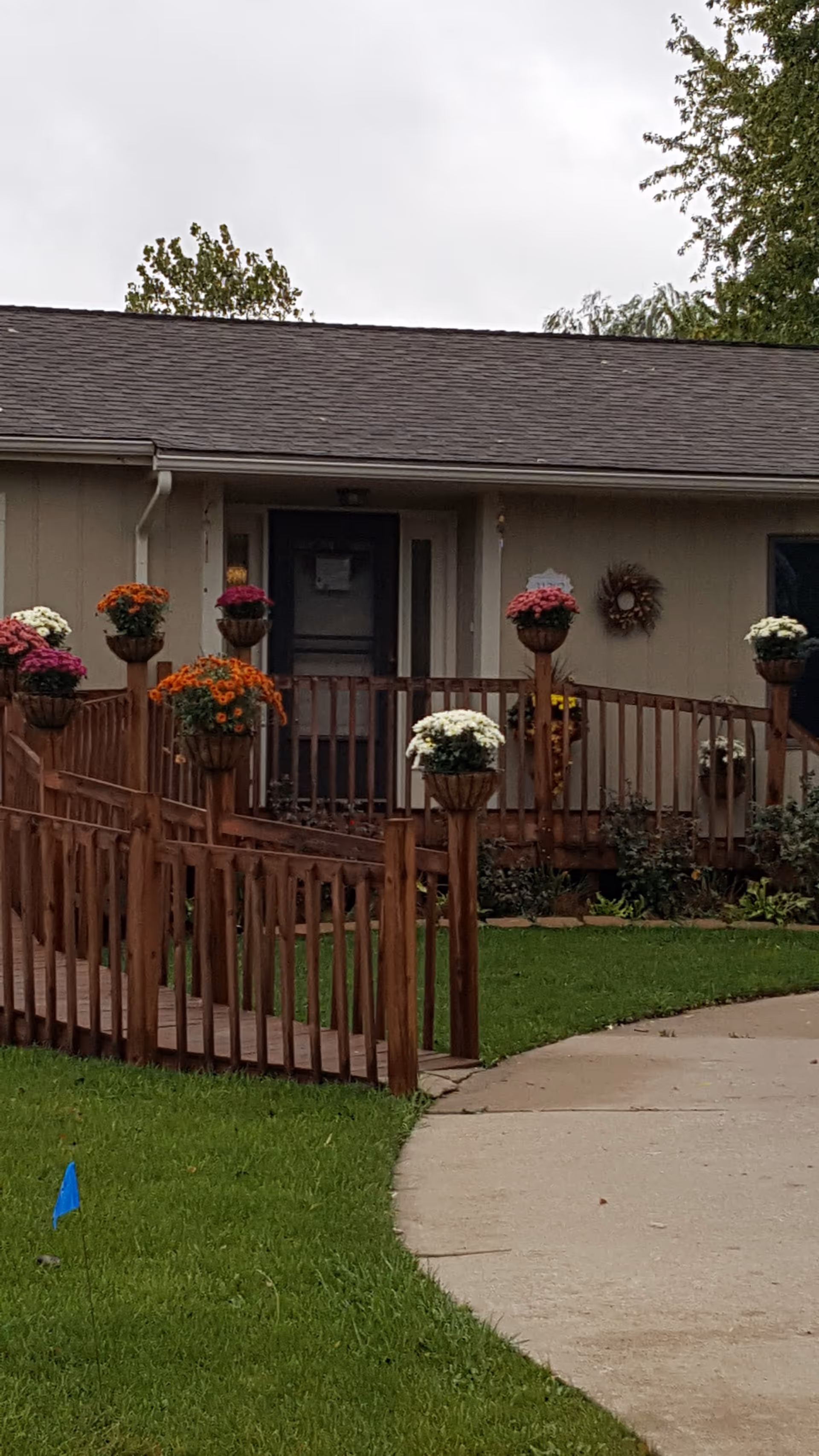 Exterior view of a single-story building with a wooden ramp leading to the entrance. The ramp is decorated with flower pots containing colorful flowers. There is a wreath hanging on the wall near the door, and green grass surrounds the walkway.