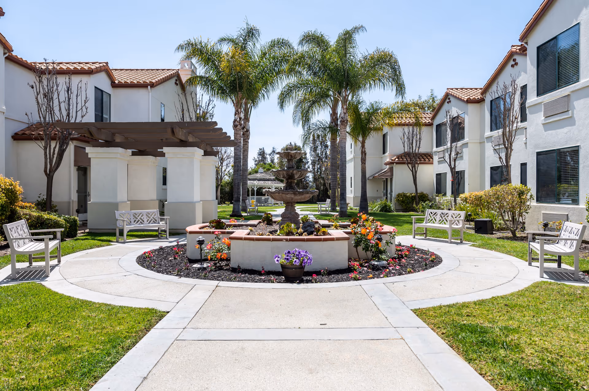 Outdoor courtyard area at Aegis Living Ventura featuring a central multi-tiered water fountain surrounded by flower beds and a circular walkway. There are white benches placed around the walkway, palm trees, and two-story white buildings with red-tiled roofs in the background under a clear blue sky.