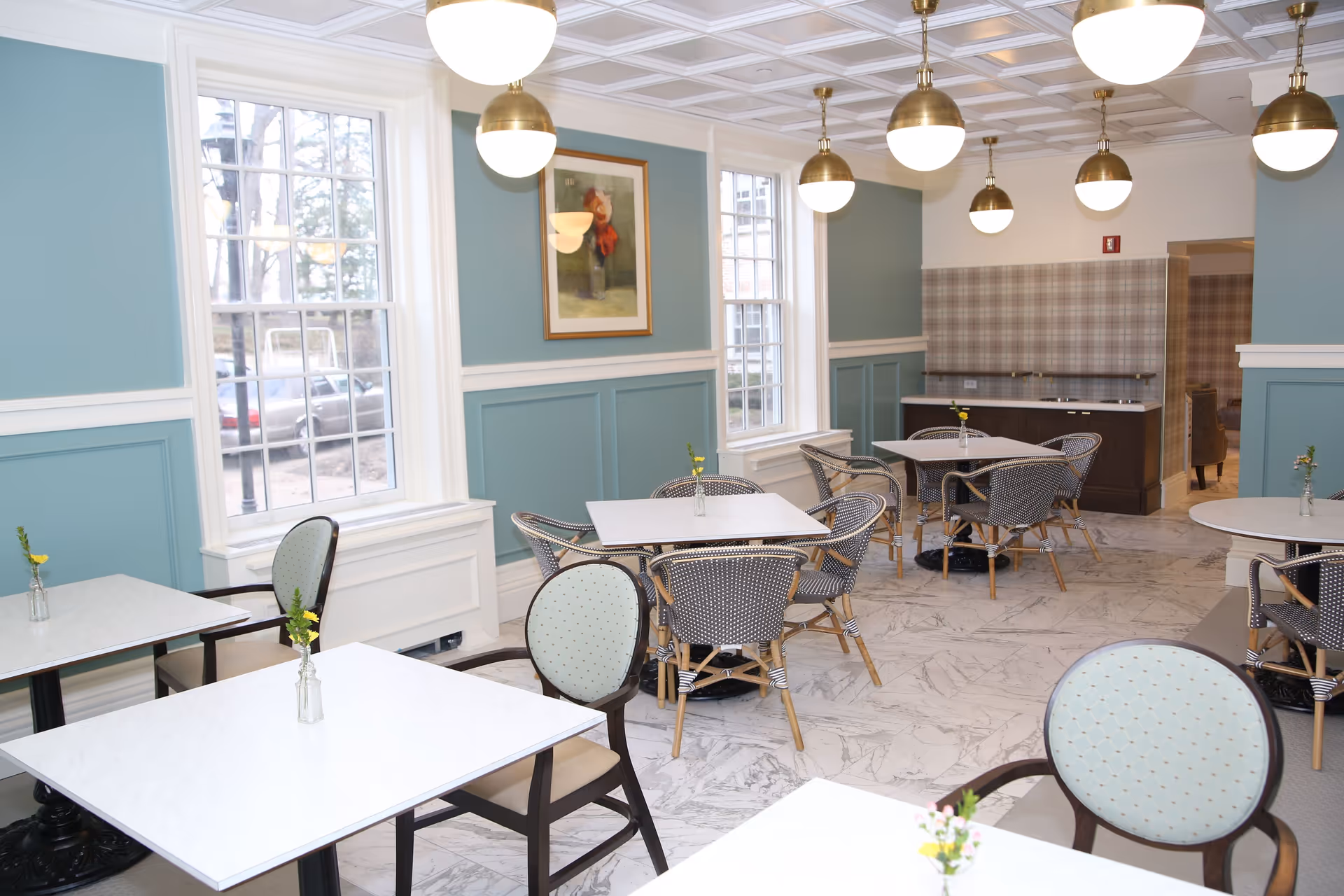 A bright dining room with white marble floors, light blue walls with white trim, and large windows letting in natural light. The room contains several square and round tables with chairs around them, each table decorated with a small vase holding yellow flowers. Brass pendant lights hang from the ceiling, and a framed painting is visible on one wall.