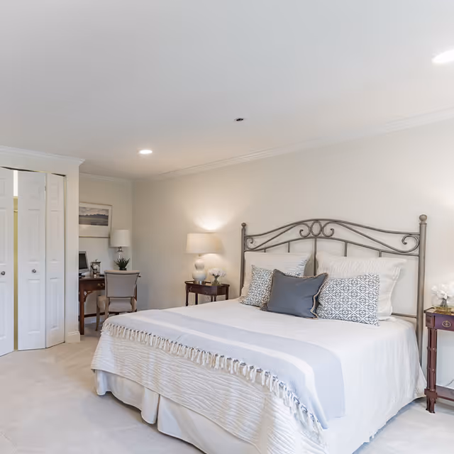 A bright and neatly arranged bedroom featuring a large bed with a decorative metal headboard, white and patterned pillows, and a light-colored bedspread with a fringed throw. On either side of the bed are small wooden nightstands with lamps and floral decorations. In the background, there is a small desk with a chair, a computer, and a framed picture on the wall. The room has light-colored walls and carpeted flooring.