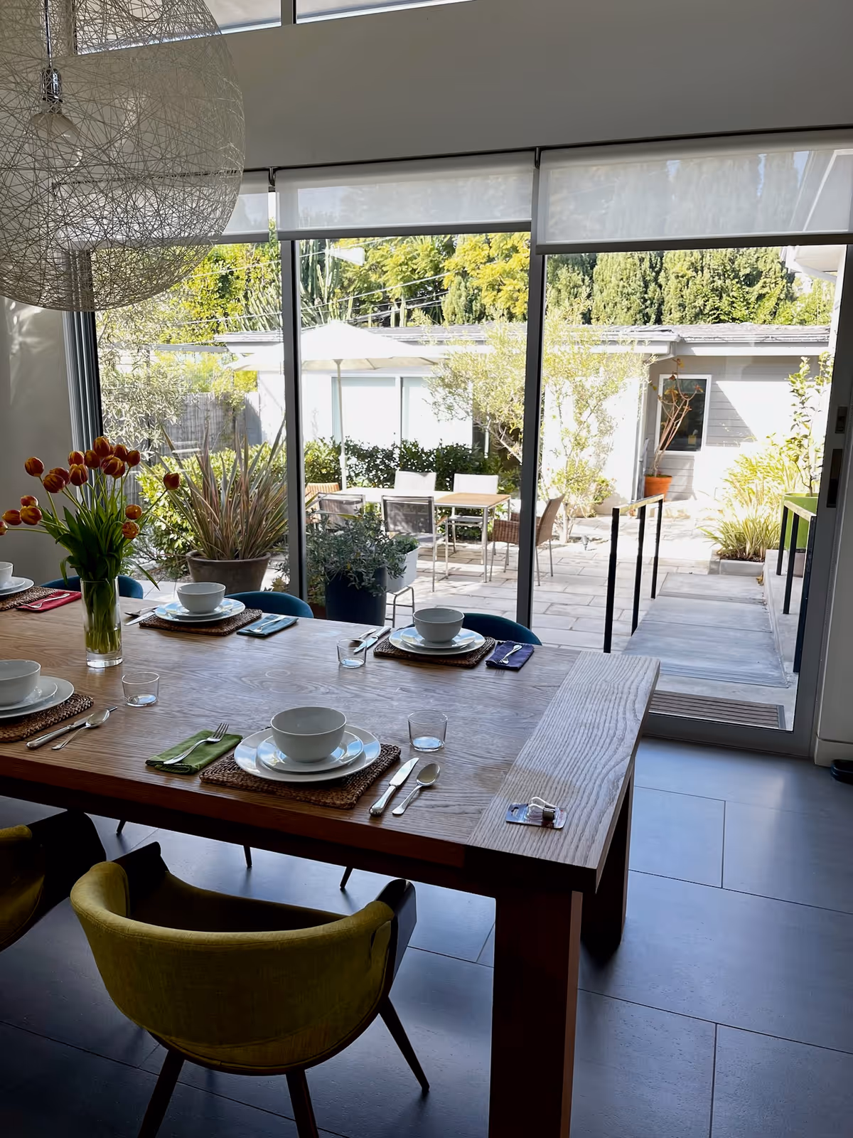 A dining area with a wooden table set for four people, each place setting including a bowl, plate, glass, and cutlery. A vase with red tulips is on the table. The room has large sliding glass doors that open to an outdoor patio with chairs, tables, and various plants. The floor is tiled, and there is a modern hanging light fixture above the table.