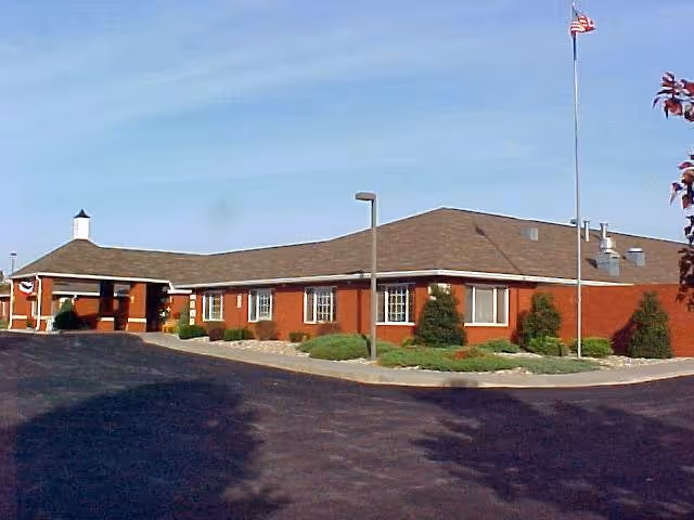 Single-story brick building with a brown shingled roof, several windows, and a covered entrance. There is a flagpole with an American flag in front of the building, surrounded by landscaped bushes and a paved driveway.