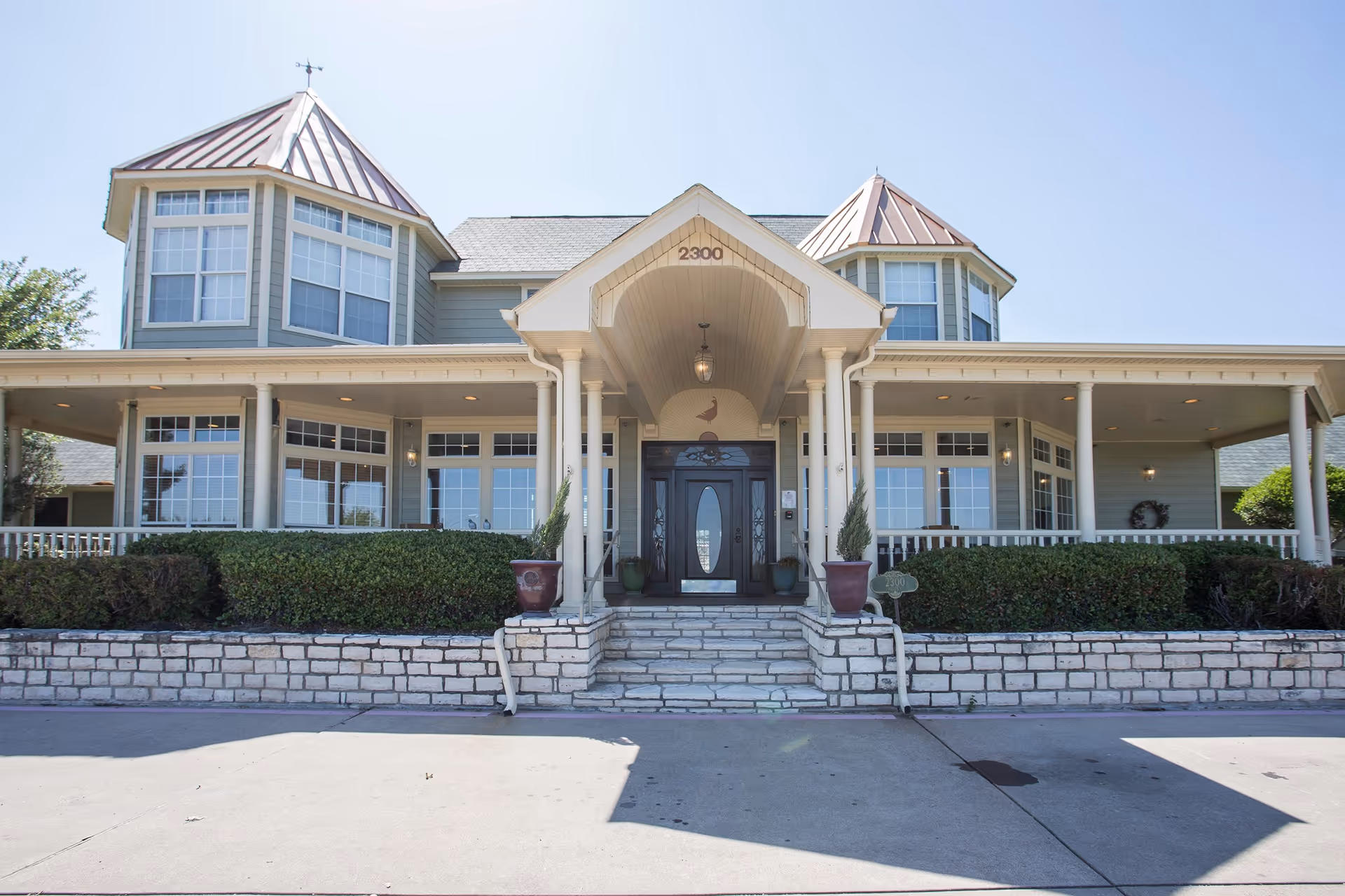 Front exterior view of a senior living facility building with a covered entrance, two large bay windows with copper roofs, and a stone staircase leading up to a dark wooden door. The building is surrounded by neatly trimmed bushes and has a clear blue sky in the background.