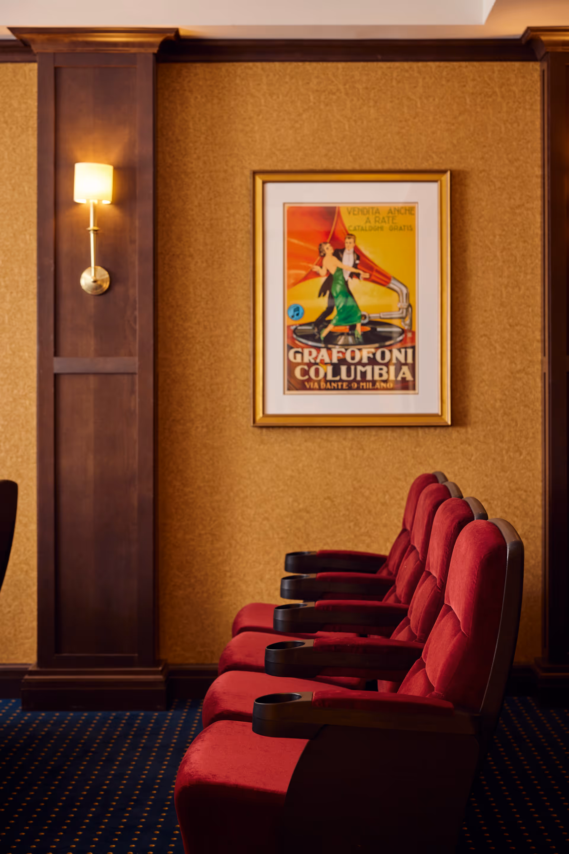 A row of red cushioned theater-style seats with wooden armrests and cup holders, positioned against a wall with brown paneling and a vintage framed poster featuring a dancing couple and a gramophone. A wall sconce with a lit lamp is mounted on the wooden panel to the left.
