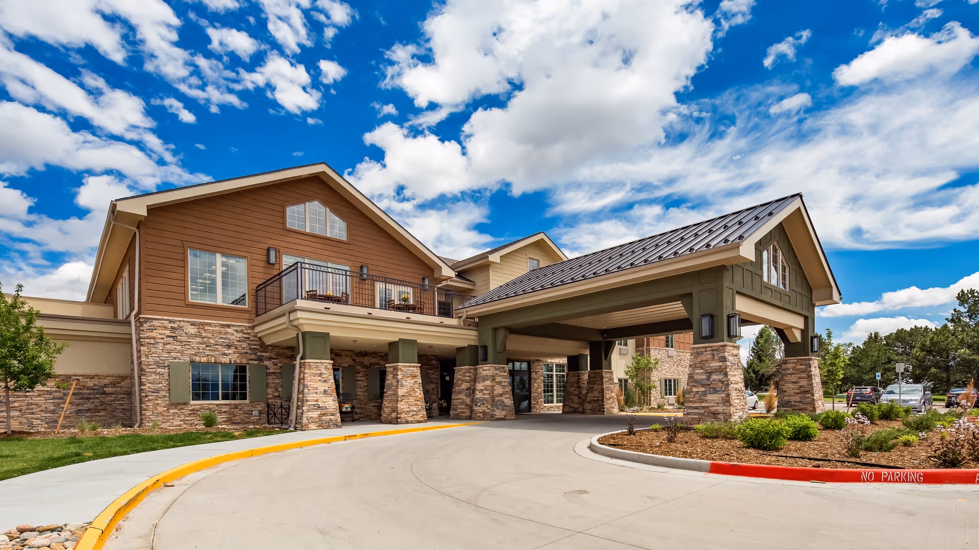 Front entrance of a senior living building with a covered porte-cochere, stone columns, balcony, and driveway under a blue sky.