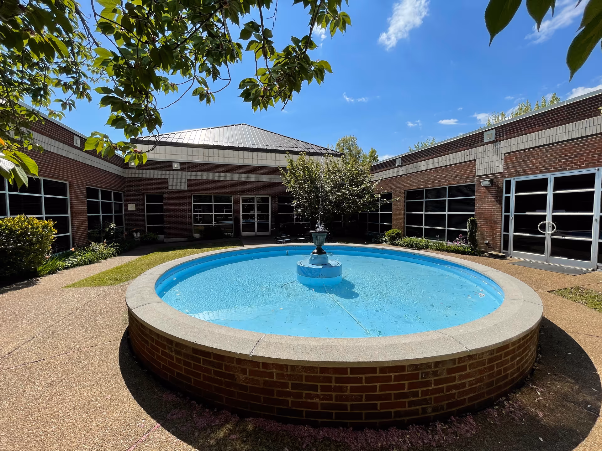 Outdoor courtyard area with a circular water fountain in the center, surrounded by a brick building with large windows and glass doors. There are trees and plants around the courtyard under a clear blue sky.