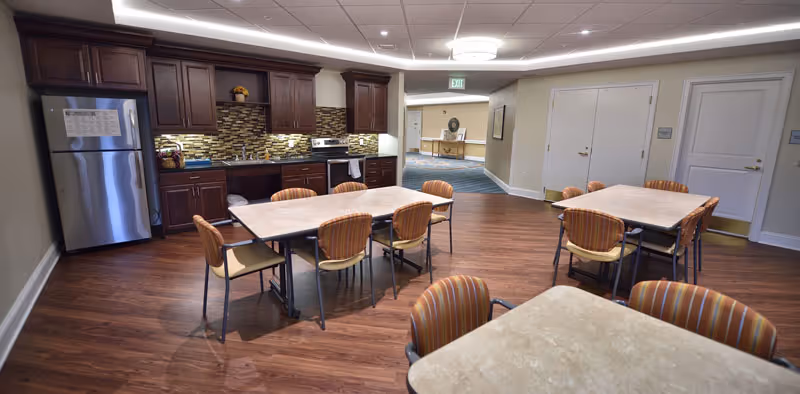 Interior view of a senior living facility kitchen and dining area with wooden flooring, dark wooden cabinets, stainless steel refrigerator, stove, and two tables each surrounded by six striped cushioned chairs. The room is well-lit with ceiling lights and has beige walls. A hallway and additional room are visible in the background.