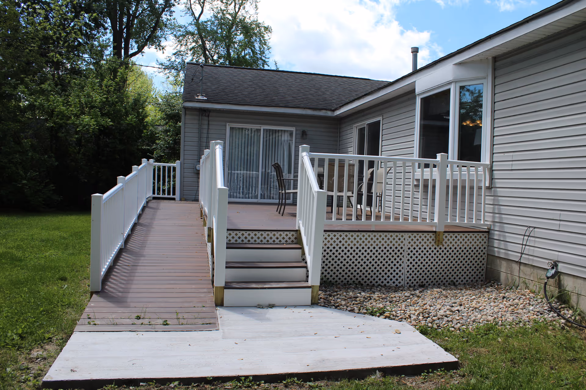 Outdoor wooden deck attached to a single-story building with white railings, a wheelchair accessible ramp, stairs, and patio furniture. The area is surrounded by green grass and trees under a partly cloudy sky.