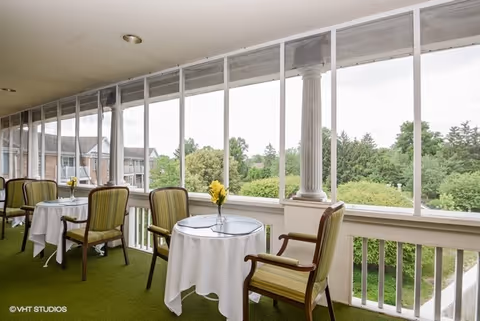 A screened-in porch area with small round tables covered with white tablecloths, each accompanied by two striped cushioned chairs. Each table has a small vase with yellow flowers. The porch overlooks a green landscape with trees and residential buildings in the background.